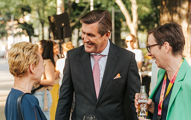 Two women and one men discussing outdoors at an event, other people in the background.